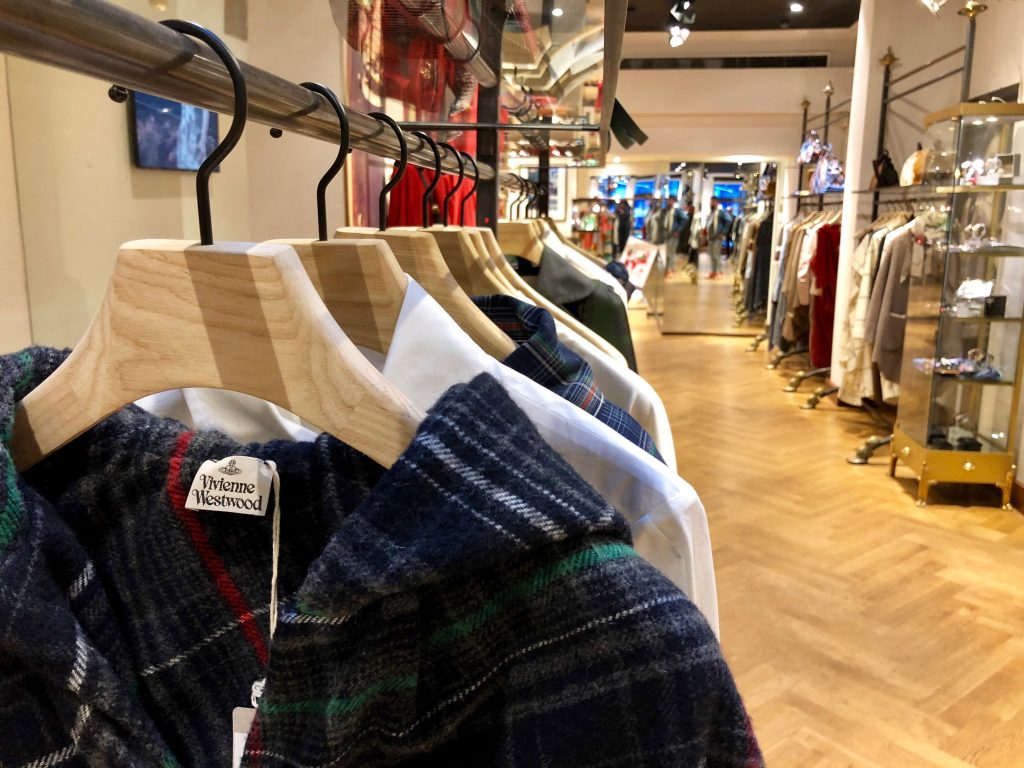 HOL Group A row of clothes on wooden hangers in a brightly lit boutique, featuring a mix of patterns and fabrics, including plaid reminiscent of Vivienne Westwood's designs. The store showcases wooden flooring and shelves along the walls displaying various items, with a long aisle leading to the back.