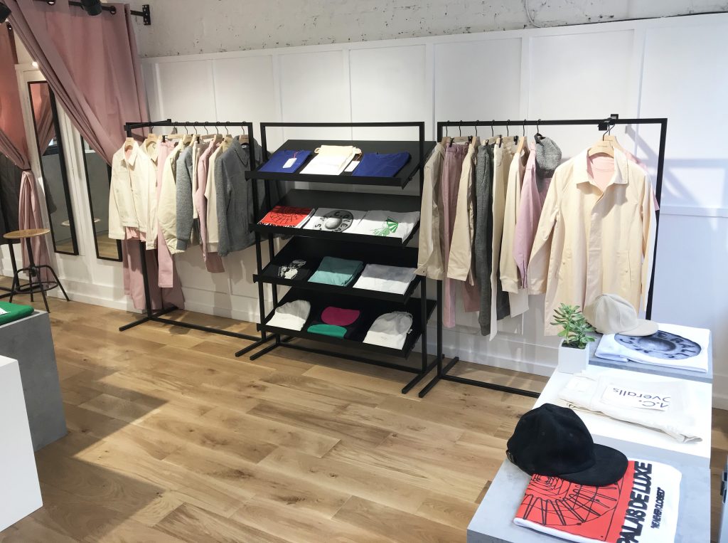 HOL Group A modern clothing store with wooden flooring. Three black racks display various clothing items like shirts, jackets, and MC Overalls in neutral and pastel colors. Folded shirts and accessories are on shelves and a table. A fitting room is visible in the background.