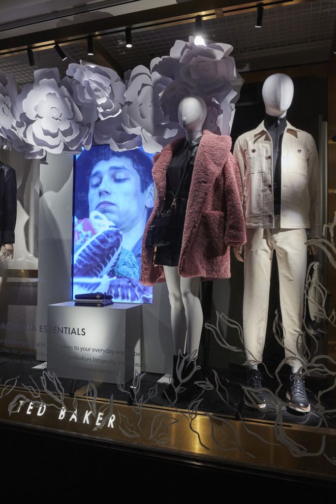 HOL Group The store window display features white floral decorations, two headless mannequins dressed in sophisticated Ted Baker white and beige outfits, and a background image of a person in a serene pose with closed eyes surrounded by white flowers. The display elegantly showcases Ted Baker clothing.