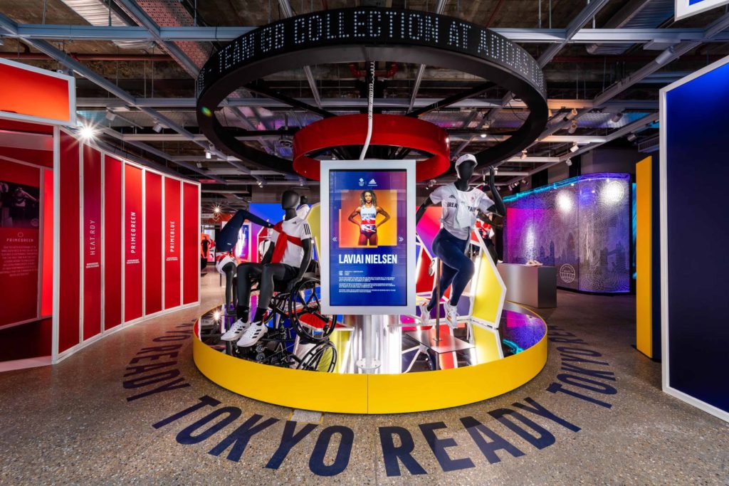 HOL Group A retail display featuring mannequins wearing athletic gear inside a vibrant, modern store nods to the Tokyo Olympics 2020. Overhead signage reads, "Move to Zero Collection at House of Innovation." A central screen displays information about an athlete, surrounded by colorful, geometric structures.