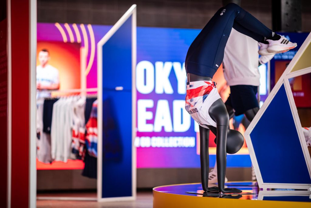 HOL Group A retail display features a mannequin in a handstand pose, dressed in athletic wear with a white, red, and blue top, and navy leggings. Behind the mannequin are clothing racks and vibrant signage proudly showcasing Tokyo 2020 apparel, creating an energetic and eye-catching scene.