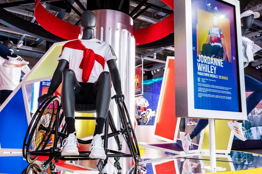 HOL Group A display features a mannequin in a wheelchair wearing white athletic attire with a red accent top. Next to it is a large poster highlighting para-athlete Jordanne Whiley, showcasing her achievements at the Tokyo 2020 Olympics and featuring her image. The background is vibrant and sporty.