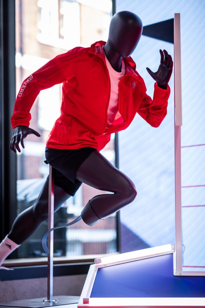 HOL Group A black mannequin dressed in a red jacket, white shirt, and black shorts is posed in a dynamic running position inside a well-lit room. There is a window in the background partially showing an urban street scene reminiscent of Tokyo during the 2020 Olympics.