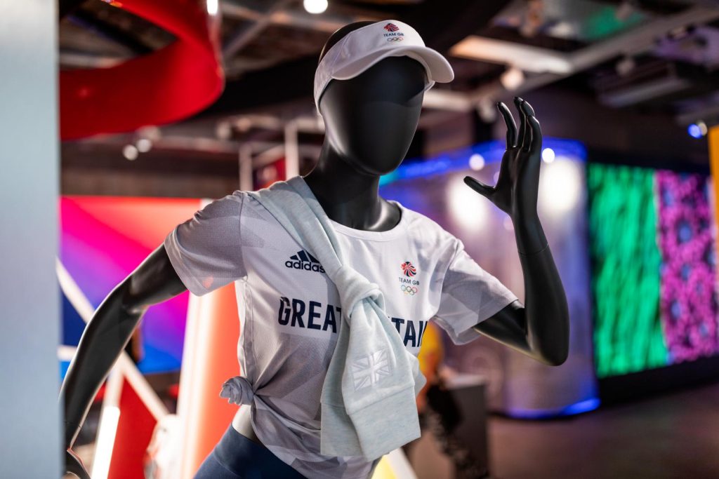 HOL Group A mannequin dressed in athletic wear featuring a "Great Britain" logo is posed as if running, reminiscent of the 2020 Tokyo Olympics. It wears a white cap, a white shirt, and has a light grey sweatshirt tied around its waist. The background is brightly lit with a colorful display.