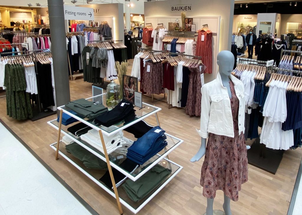 HOL Group A clothing store with various racks displaying neatly arranged women's clothes. A mannequin in the foreground is dressed in a patterned dress and white jacket from Baukjen. Signs for fitting rooms are visible, while shoppers are seen browsing in the background.