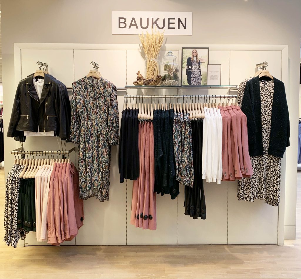 HOL Group A Baukjen clothing display showcases a chic black leather jacket, patterned dresses, and diverse colored skirts and tops in black, white, and pink hues. The elegant setup is enhanced by a prominent Baukjen sign at the top, with decorative dried plants and a framed photo adding a sophisticated touch.