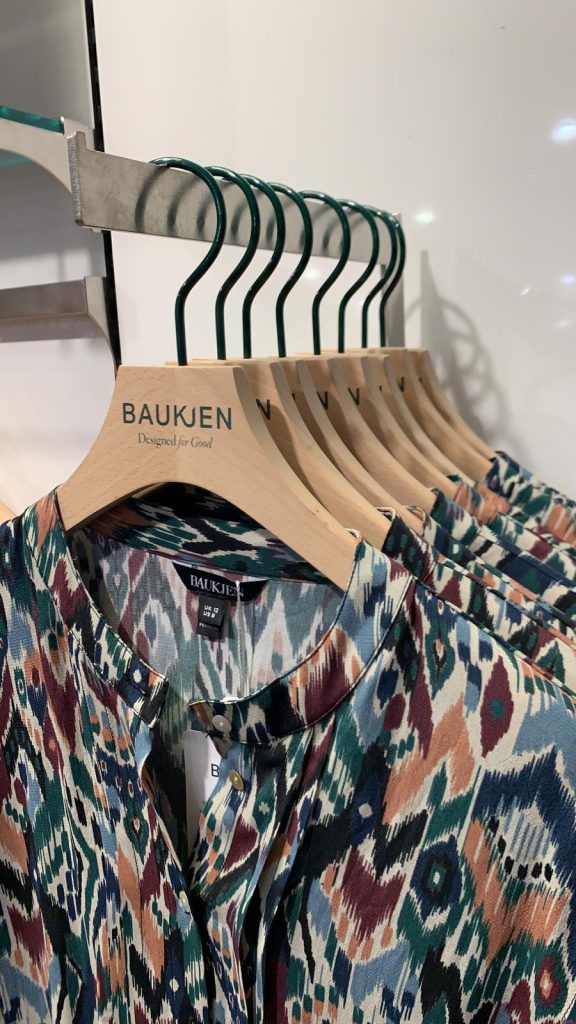 HOL Group A row of Baukjen hangers displaying colorful, patterned blouses with intricate designs in shades of blue, green, brown, and white. The metal hangers with wooden tops are neatly arranged on a metal rod.