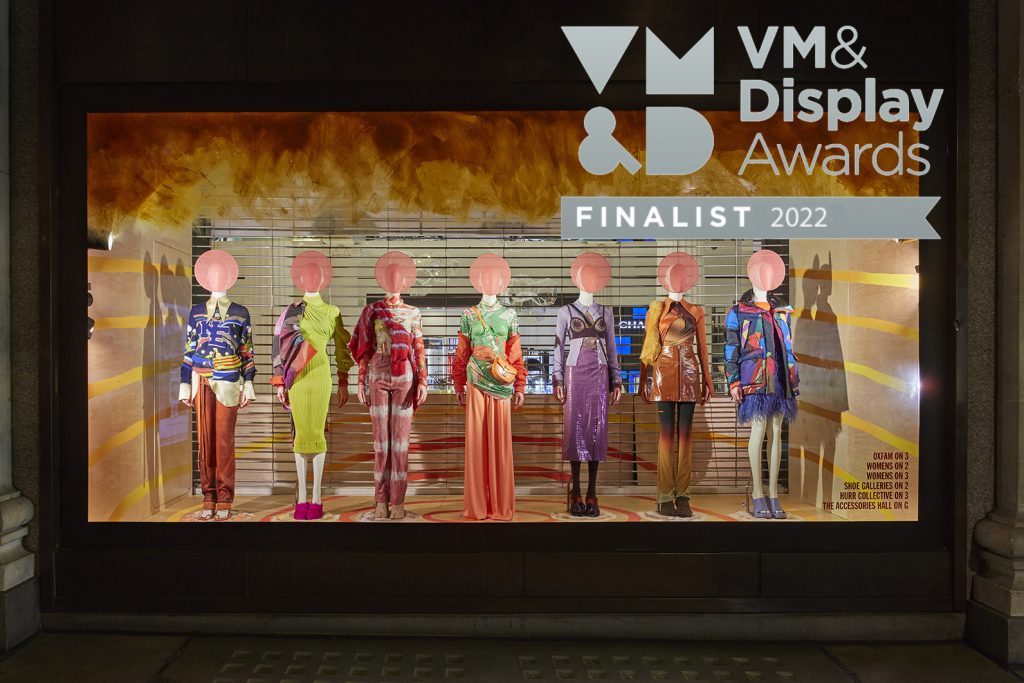 HOL Group A retail store window displays six mannequins wearing various colorful and stylish outfits. The window features a sign that reads "VM& Display Awards FINALIST 2022." The mannequins have spherical, abstract heads, and the background has a warm, gradient pattern, reminiscent of Selfridges' luxury shopping ambiance.