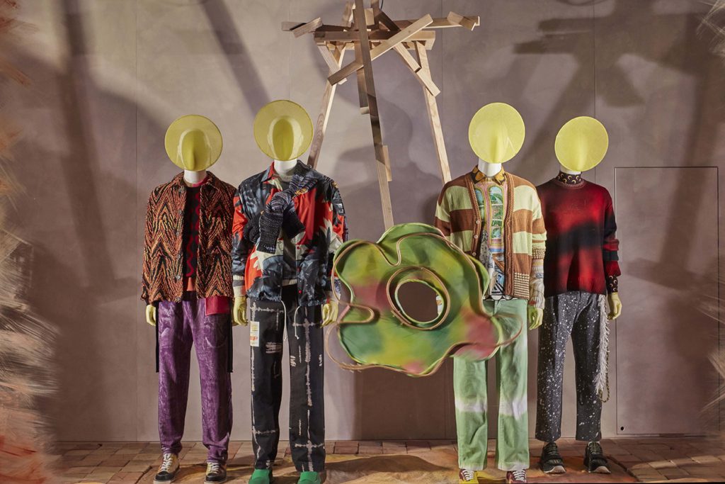 HOL Group Four mannequins dressed in eclectic, colorful clothing stand side by side inside Selfridges. Each mannequin has a circular yellow disk instead of a face. Behind them, a wooden structure and a large abstract green and yellow shape add to the artistic display in this luxury shopping destination.