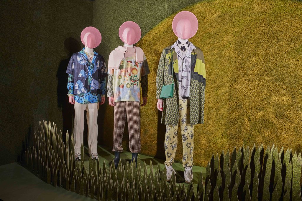 HOL Group Three mannequins dressed in colorful, eclectic outfits stand in a grassy display at Selfridges. They wear large, pink circular hats that obscure their faces. The backdrop features green and yellow hues, resembling a sun setting behind rolling hills, evoking the charm of high-end shopping at this luxury department store.