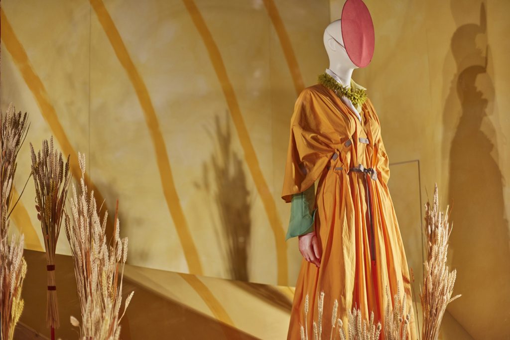 HOL Group A white mannequin at Selfridges wears a flowing orange dress with a green inner layer and a wide belt. It has a red, circular face covering and stands amidst dry wheat stalks. The background features a warm yellow wall with artistic shadows and patterns, embodying the essence of luxury retail.