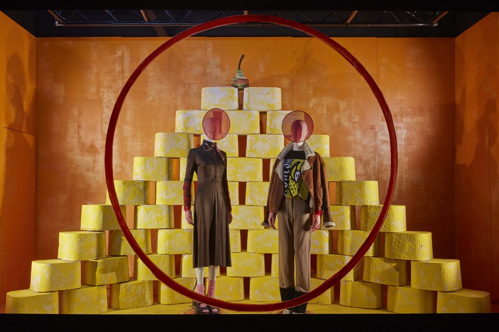 HOL Group Two abstract mannequins with circular cutouts for heads stand within a large red circle in front of a pyramid made of yellow blocks. One mannequin wears a brown dress and the other dons a checkered suit with a green and yellow design, reminiscent of the avant-garde displays at Selfridges department store.