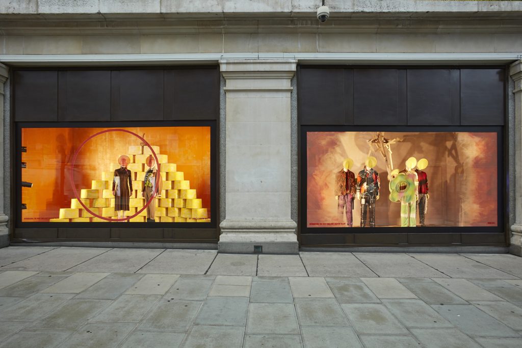 HOL Group A luxury department store's storefront window display features mannequins with brightly colored outfits. The left window displays two mannequins in front of a yellow pyramid backdrop, while the right window has three mannequins wearing high-end fashion and props against an orange background.