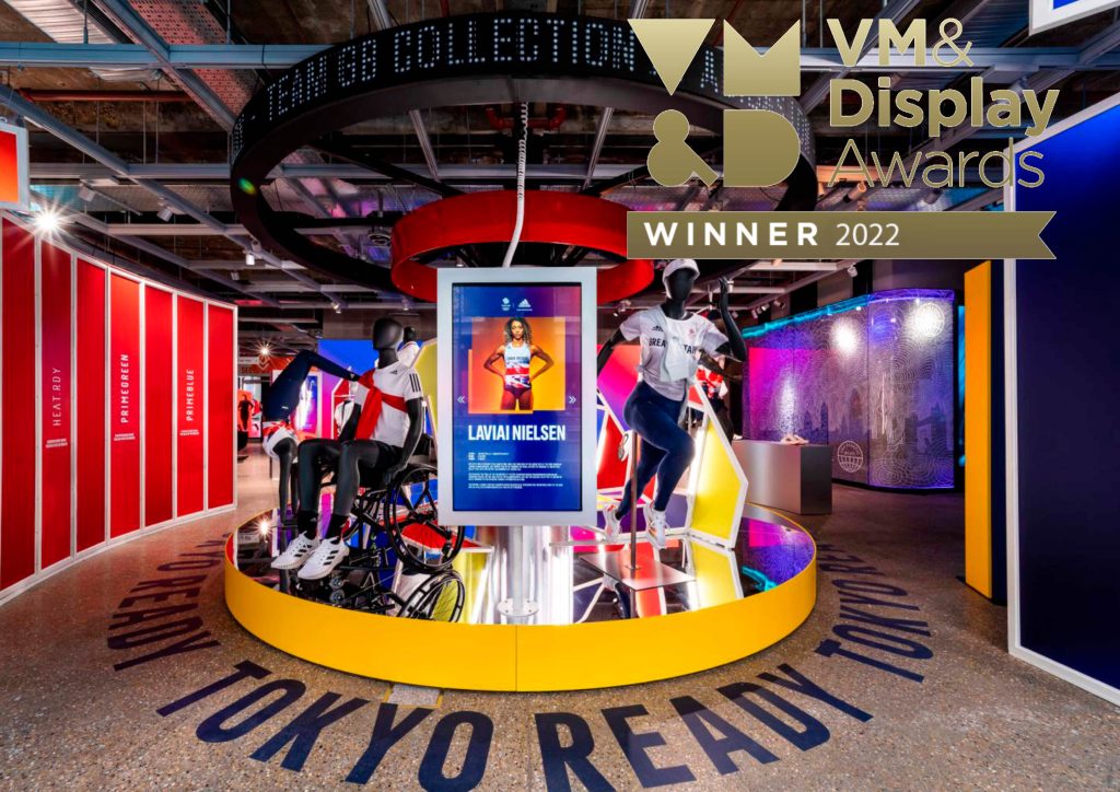 HOL Group A vibrant display with mannequins wearing athletic clothing stands inside a store adorned with red, yellow, and blue panels. A central screen showcases an athlete from the Tokyo 2020 Olympics. The image features a 2022 VM & Display Awards winner banner at the top right corner.