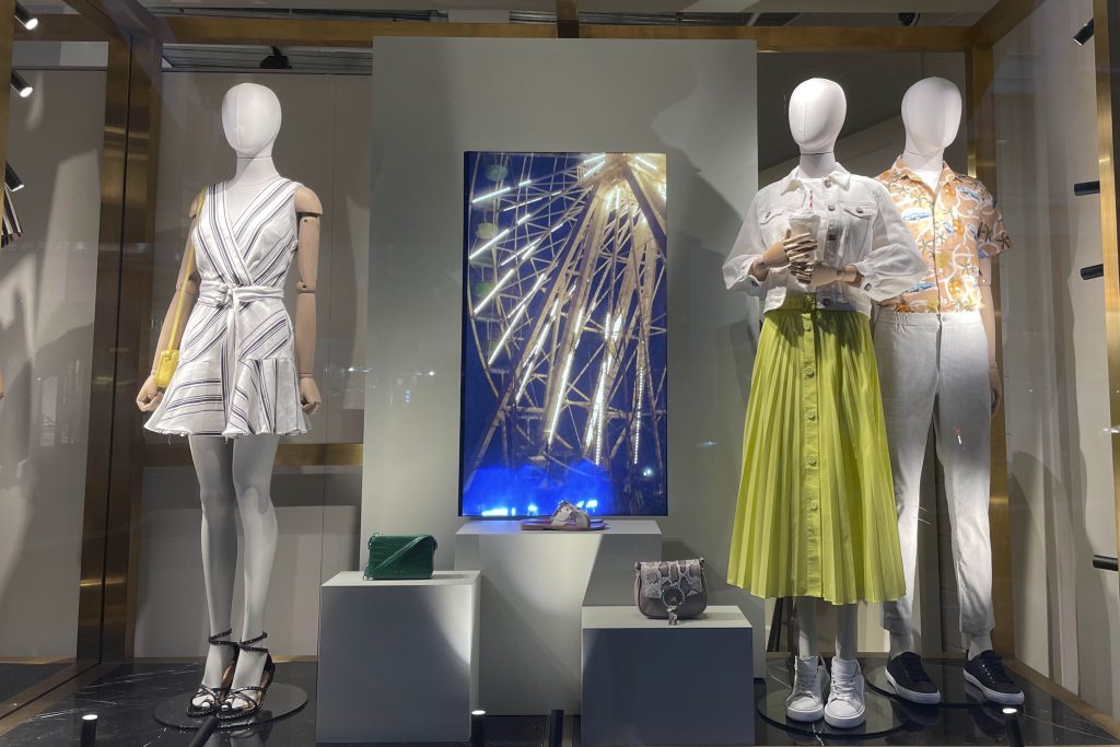 HOL Group A store window display features three mannequins dressed in trendy Ted Baker outfits. The center backdrop showcases an illuminated Ferris wheel. The left mannequin wears a striped dress, the middle one a casual top with a green skirt, and the right one a white shirt and gray pants.
