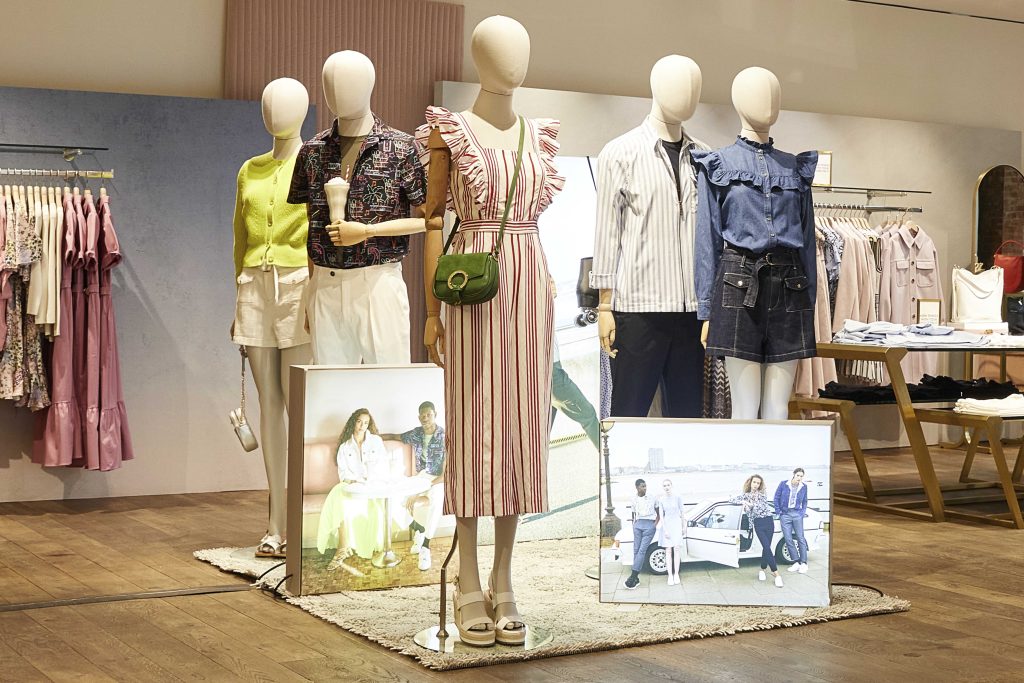 HOL Group A Ted Baker clothing store display features mannequins dressed in various summer outfits, including dresses and casual wear. The background includes a rack of clothes and two framed photos of people posing with a car. The display is set on a wooden floor.