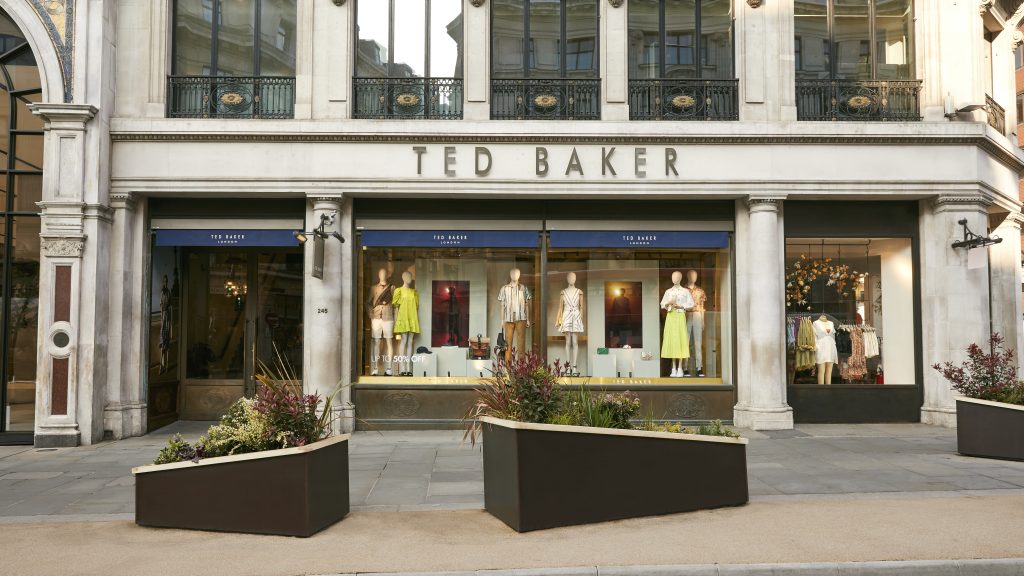 HOL Group A store facade of the Ted Baker clothing brand, with large glass windows displaying mannequins dressed in various outfits. The storefront has a blue awning emblazoned with "Ted Baker" and is framed by elegant architectural details. Planters with greenery are placed on the sidewalk in front.