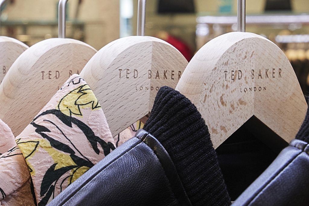 HOL Group A close-up shot of Ted Baker branded wooden coat hangers with various clothing items on them. Some garments feature floral patterns, while others are plain, all hanging on a metal rack in a Ted Baker retail store.