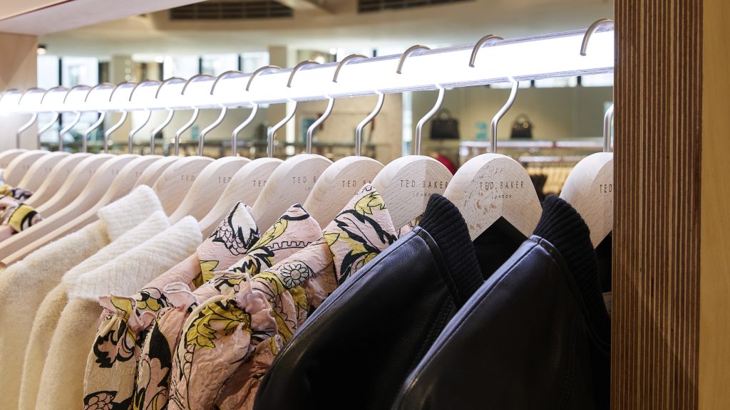 HOL Group A row of Ted Baker clothing items on hangers, including a mix of patterned jackets and solid-colored garments, displayed on a wooden rack under soft lighting. The hangers are branded with "Ted Baker." The background shows an out-of-focus retail store setting.