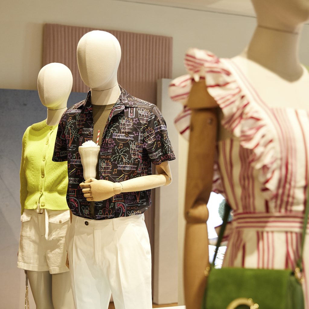 HOL Group Three mannequins are dressed in summer attire. The front mannequin wears a pink and white striped dress with ruffled sleeves and a green purse. The middle mannequin sports a Ted Baker green sweater, white shorts, and holds an ice cream cone. The back mannequin wears a dark shirt with white pants.