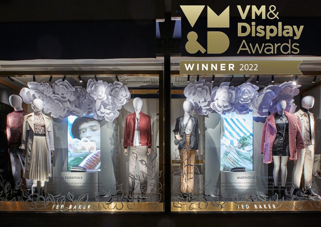 HOL Group A store display window showcasing mannequins dressed in stylish Ted Baker outfits. The display is adorned with large white paper flowers and features screens with photos of nature. "VM & Display Awards Winner 2022" is displayed at the top right, with the brand "Ted Baker" elegantly noted at the bottom.