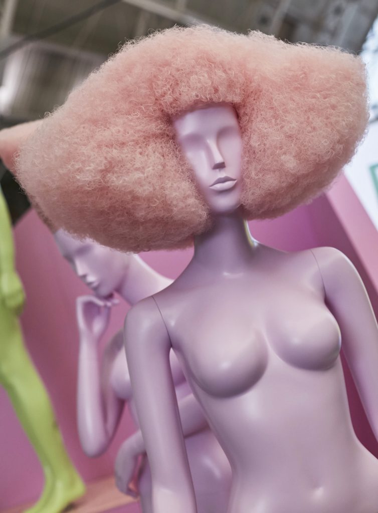 HOL Group A lavender-colored mannequin with a tall, voluminous pink afro hairstyle stands in the foreground at the VM & Display Show 2022. Another lavender mannequin sits behind it, hand resting on their chin. The background includes pastel geometric shapes and soft lighting.