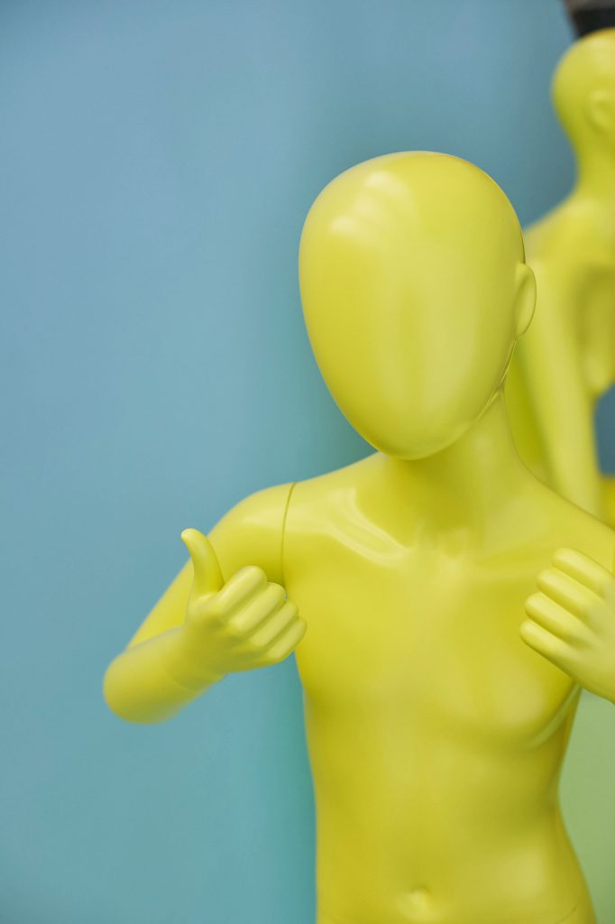 HOL Group A yellow faceless mannequin with a smooth surface is posed against a blue background, giving a thumbs-up with both hands. Another identical mannequin is partially visible in the background.
