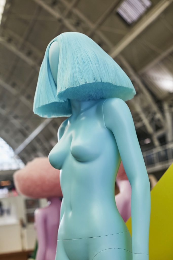 HOL Group A blue mannequin with a wig-like hairstyle stands indoors, seemingly at an art exhibition. The setting features industrial architectural elements, including large windows and high ceilings. Another mannequin can be seen blurred in the background, making this VM display a highlight of Show 2022.