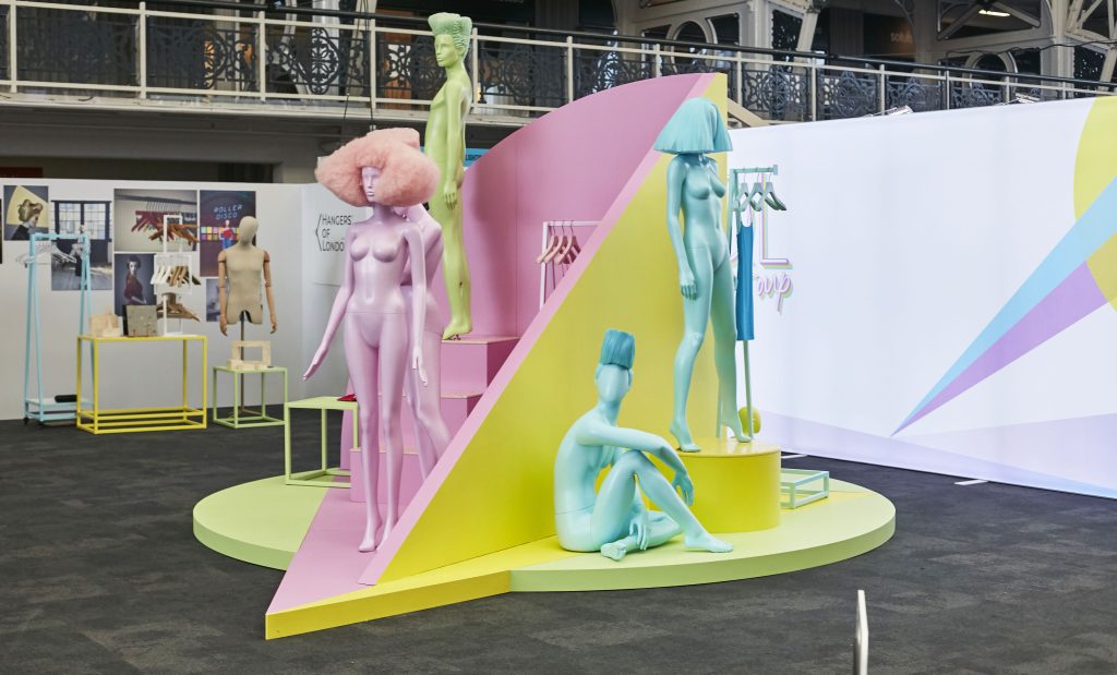 HOL Group A vibrant art installation from 2022 features pastel-colored mannequins with quirky hairstyles posed on a geometric, multicolored platform. This unique display show includes standing mannequins and one seated, all within an indoor exhibition setting accompanied by other intriguing VM displays.