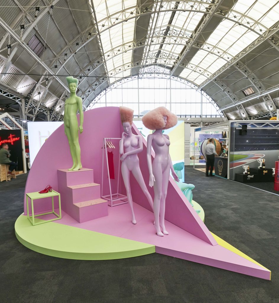 HOL Group A 2022 modern art display features purple mannequins with abstract hairstyles: one wearing a tall spiky green wig, another with a fluffy pink afro. They stand on a pink and green geometric platform under an ornate arched metal and glass ceiling, making for an eye-catching VM showcase.