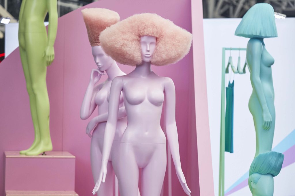 HOL Group Three pastel-colored humanoid mannequins with unique hairstyles are displayed at the 2022 VM & Display Show. The front two, pink and purple, have large, fluffy hair, while a green mannequin in the back has a modern, angled bob. One green leg and hip can be seen to the right.