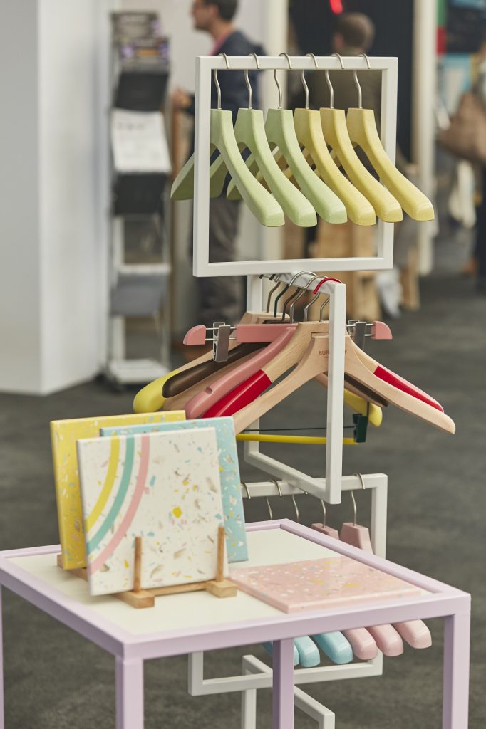 HOL Group A display in a store features colorful coat hangers on a white rack. Below, several patterned ceramic tiles are displayed on a pink table. The background shows other store fixtures and people blurred out of focus. The scene is well-lit and modern.