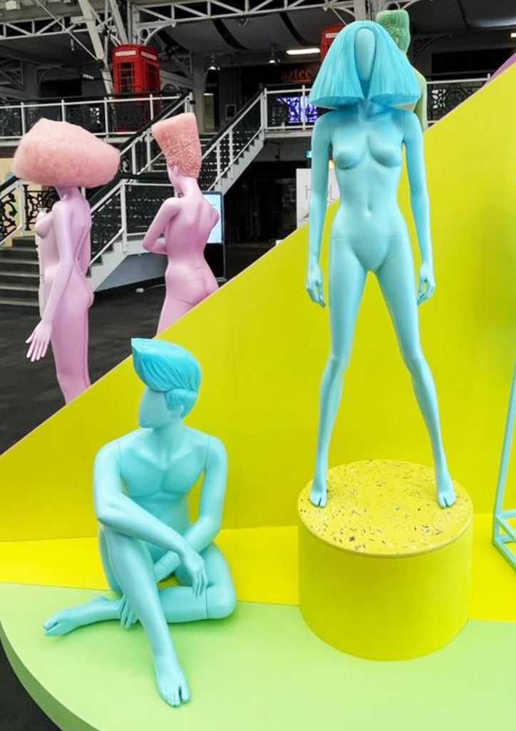 HOL Group An art installation features abstract humanoid figures in pastel colors against a yellow geometric backdrop. One blue figure stands on a pedestal with a fan-shaped headpiece, while another blue figure with a smooth head in a sitting pose is on the ground nearby.