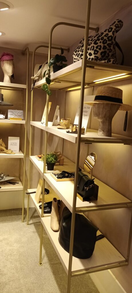 HOL Group A well-lit boutique display featuring a stunning gold, multi-tiered shelving unit by Phase Eight. Showcased are various high-end accessories including handbags, hats, and small potted plants. The items on display range in color and style, adding a chic and elegant ambiance to the space.