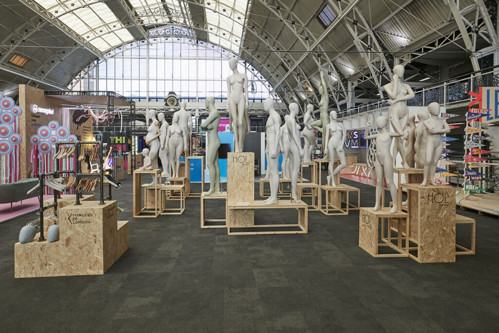 HOL Group A large indoor display at the 2023 VM & Display Show features numerous white mannequins of different sizes and poses, arranged on wooden platforms. Brightly colored art pieces and signs are visible in the background, and the area is lit by natural light streaming through a high, arched ceiling.