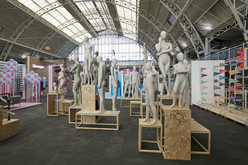 HOL Group An exhibition hall with mannequins on wooden platforms in the center, some grouped and posed artistically. Bright lighting from the high ceiling windows, various installations and displays can be seen in the background, including flags and target graphics from the 2023 VM & Display Show.