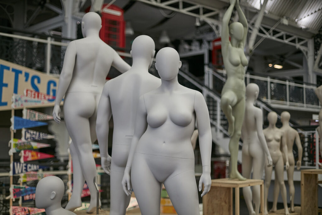 HOL Group A group of featureless mannequins, including male and female forms, are arranged in various poses in an indoor space for the 2023 Display Show. Some are standing on wooden platforms, while others are posed dynamically. The background includes industrial elements and signage, showcasing contemporary VM techniques.