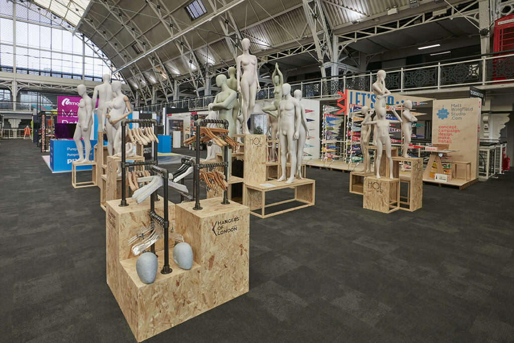 HOL Group A modern exhibition hall in 2023 features multiple mannequins without heads, some posed on wooden pedestals. Various stands display colorful clothing and accessories. The space has an industrial aesthetic with high ceilings, metal beams, and large windows in the background, creating a striking VM display show.