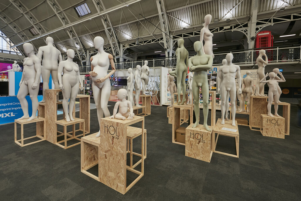 HOL Group A captivating Display Show featuring numerous mannequins, including adults and a child, is arranged on various wooden crates inside a large, well-lit exhibition hall for VM 2023. The mannequins, striking different poses and heights, are showcased on platforms marked with the letters "HOL.
