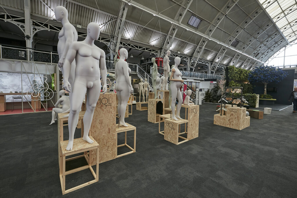 HOL Group A large indoor space at the 2023 VM & Display Show features several white, faceless mannequins in various poses, positioned on wooden crates and platforms. The room has a high, arched ceiling with metal beams and some informational displays and greenery in the background.