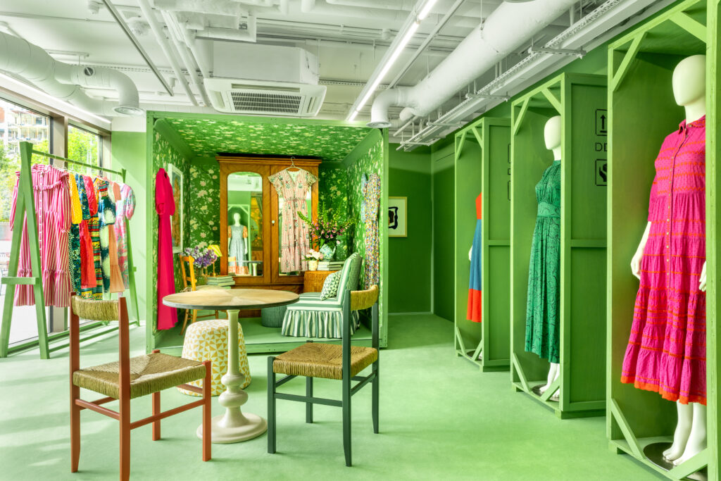 HOL Group A chic boutique interior features mannequins dressed in colorful, stylish dresses. The space is vibrant with green walls and flooring. A small sitting area with wooden chairs and a round table is in the center. Clothes hang on racks and a large mirror reflects a flower-patterned dress.