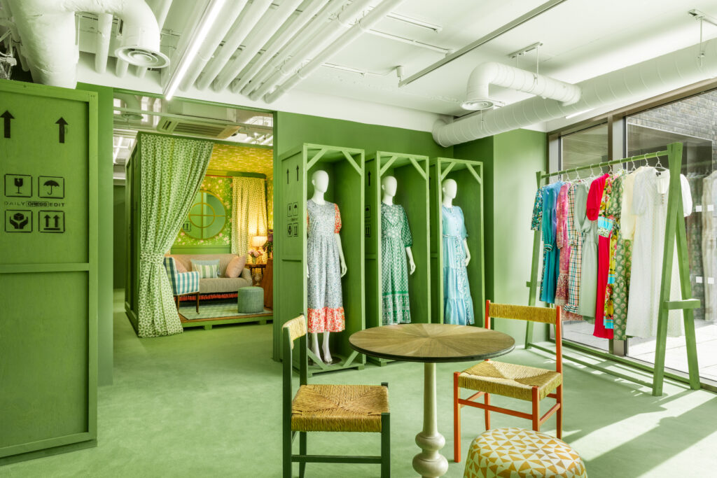 HOL Group A brightly lit, green-themed boutique displays mannequins dressed in colorful garments. The room features green walls, industrial ceiling pipes, a round table with chairs, and a cozy seating area in the background with a patterned green curtain.