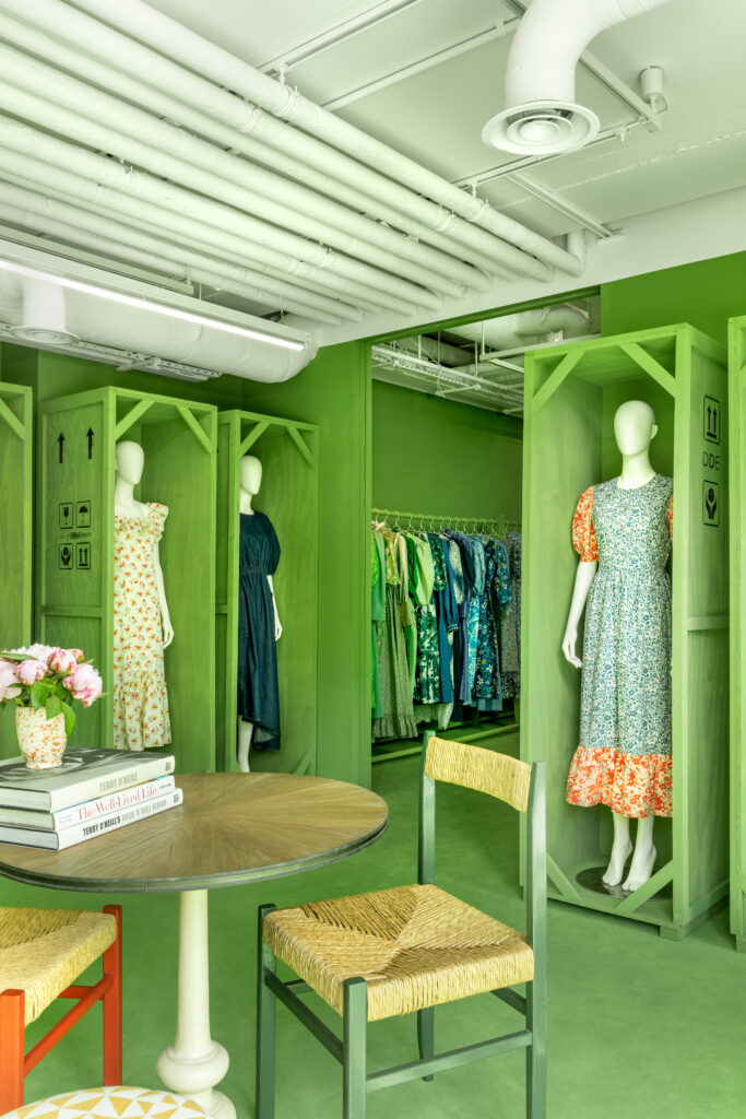 HOL Group A brightly lit room with green walls features mannequins dressed in colorful dresses. Clothes in various shades of green hang on a rack in the background. A round table with books and flowers, and two green-seated chairs, are positioned in the foreground.