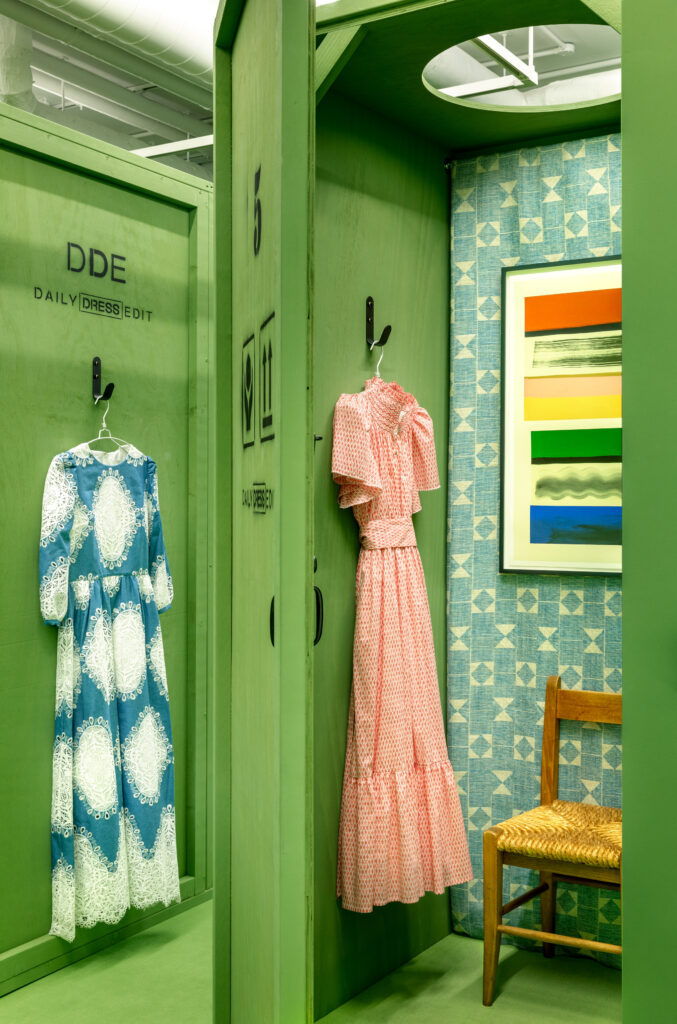 HOL Group A green dressing room with two dresses hanging: one blue with white lace and the other red polka-dotted with ruffles. A wicker chair is on the right near a blue patterned wall with a colorful framed artwork. A "Daily Dress Edit" sign is visible.