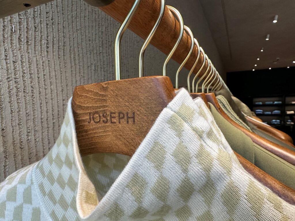 HOL Group Close-up of a rack of hangers with "Joseph" engraved on them, holding patterned knit garments in light green and cream colors. The background reveals the dimly lit interior of a Joseph clothing store.