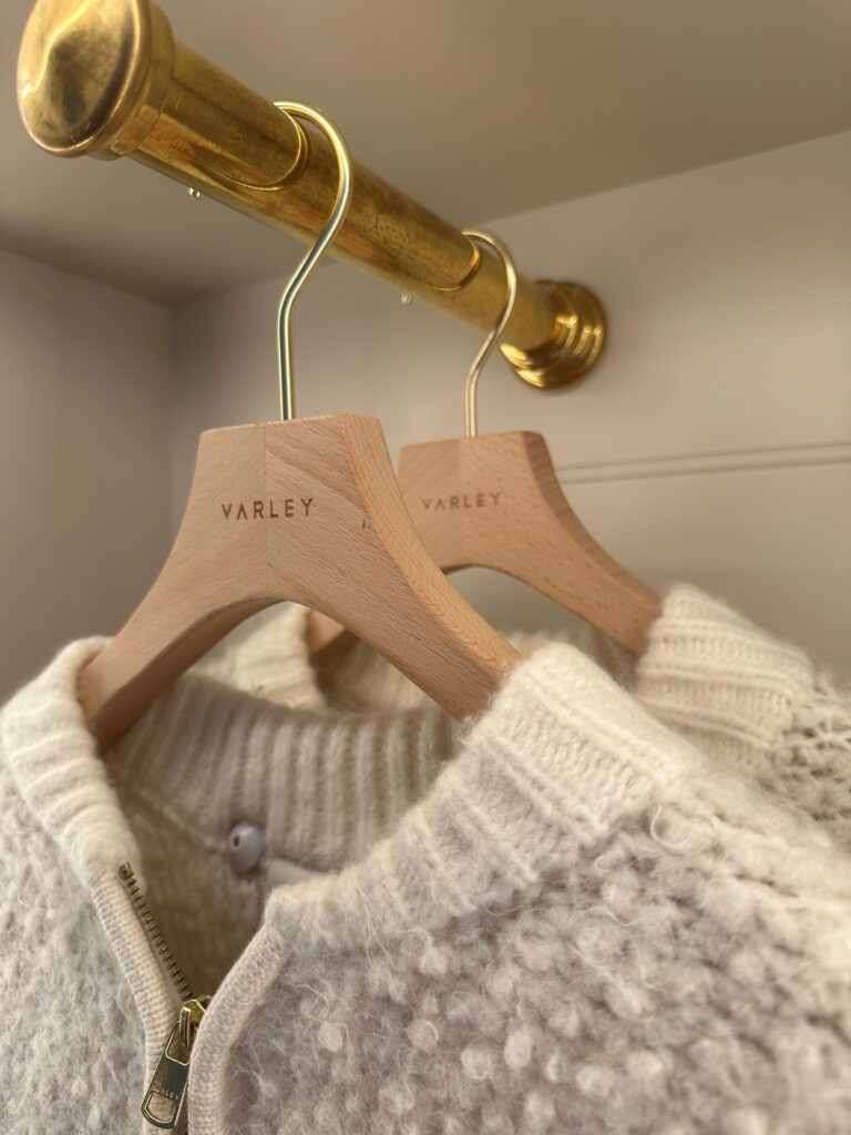 HOL Group Close-up of two Varley wooden hangers, each holding a white knit garment with a zip-up design and textured pattern on a golden rod. The brand name "Varley" is engraved on the hangers. The light neutral background enhances the cozy and organized aesthetic.