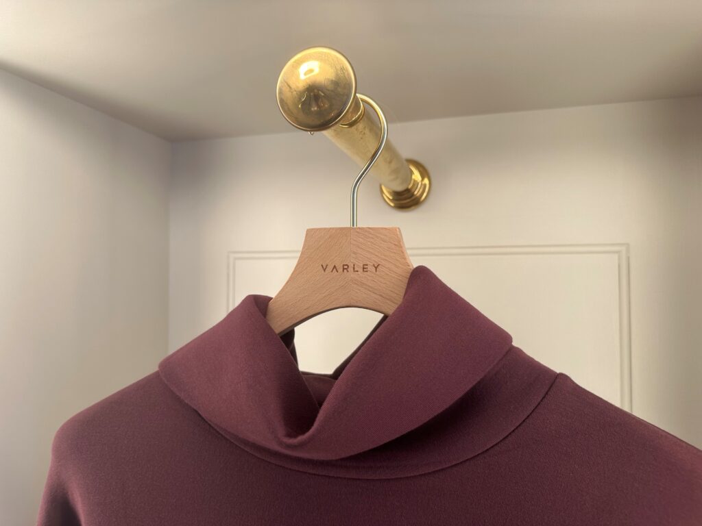 HOL Group A plum-colored top on a wooden hanger is hanging from a brass rod against a light-colored wall. The Varley-branded wooden hanger showcases the elegant garment, which features a high, folded neckline.