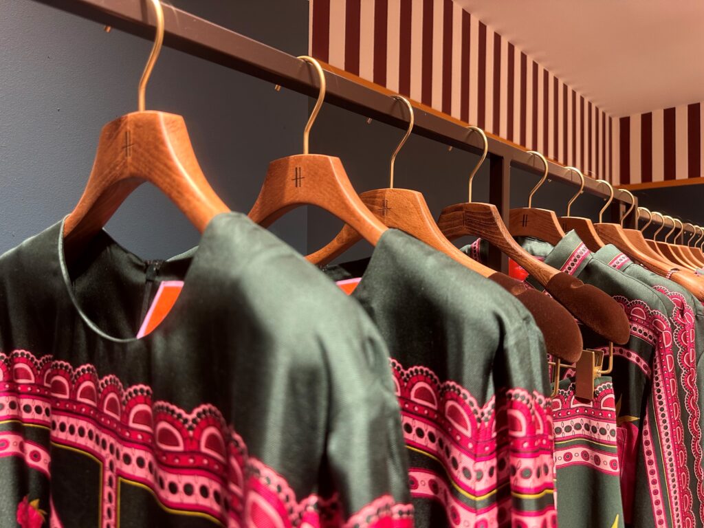 HOL Group A row of wooden hangers on a metal rod holds several identical black garments with intricate pink and red patterns. The background features a striped ceiling and dark walls, creating an elegant and coordinated display reminiscent of a luxury department store like Harrods, perfect for an elevated shopping experience.