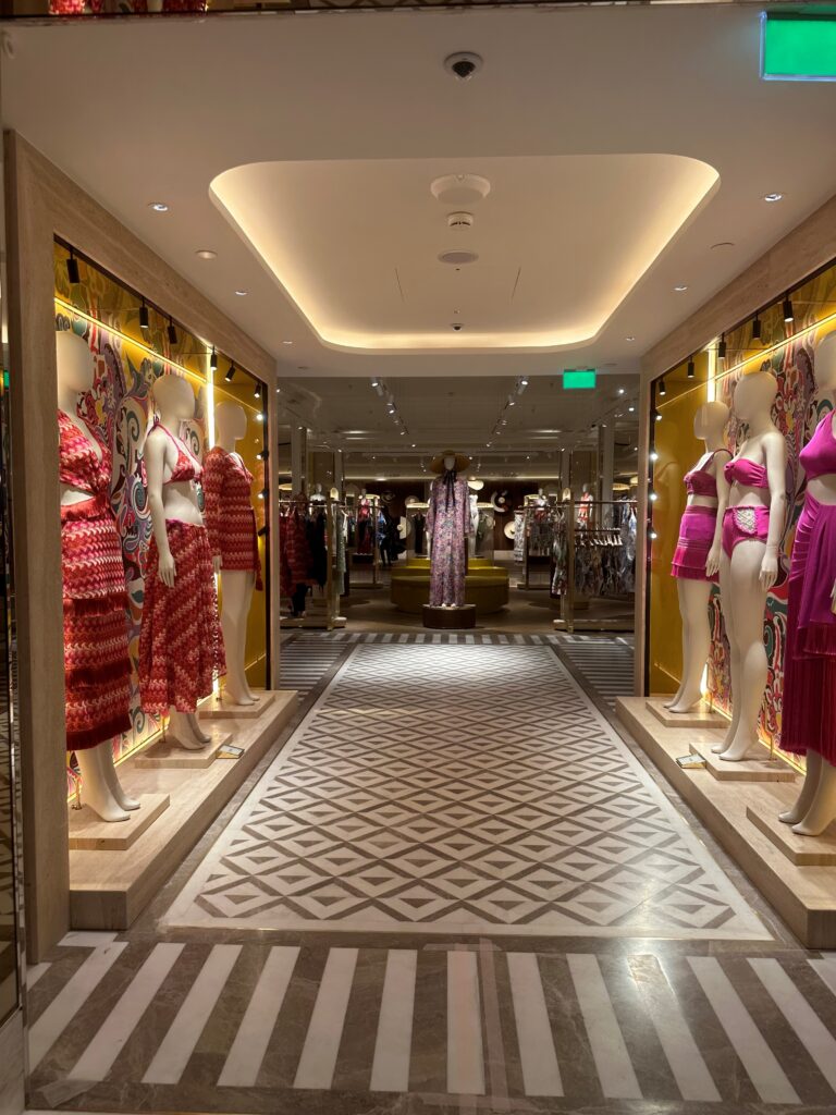 HOL Group A brightly lit boutique within Harrods features mannequins displaying colorful dresses and outfits in red and pink hues. The store has a patterned white and grey floor, with mirrors and additional clothing racks visible in the background, reflecting the elegance of a luxury department store.