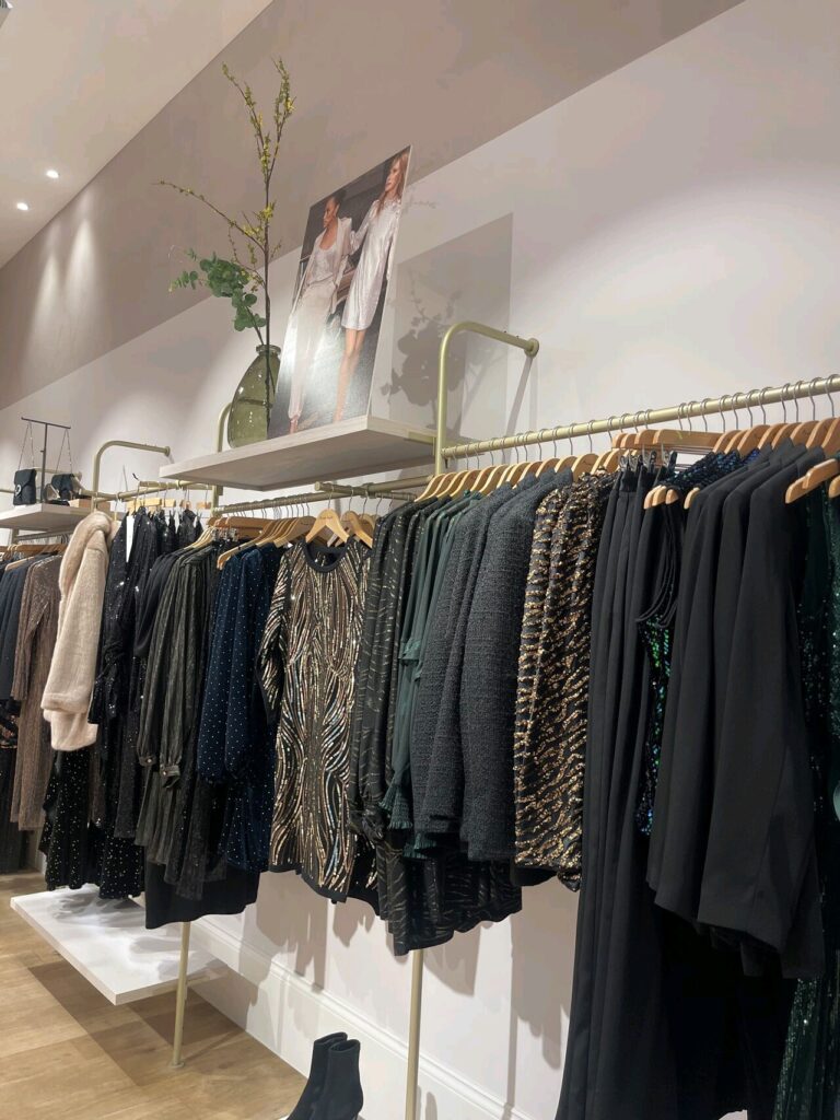 HOL Group A Phase Eight clothing store display features a variety of women's apparel on hangers. The clothes include black and brown patterned dresses, blouses, and outerwear. Above the racks, plants and a poster of fashion models adorn the shelves. The room has bright lighting.